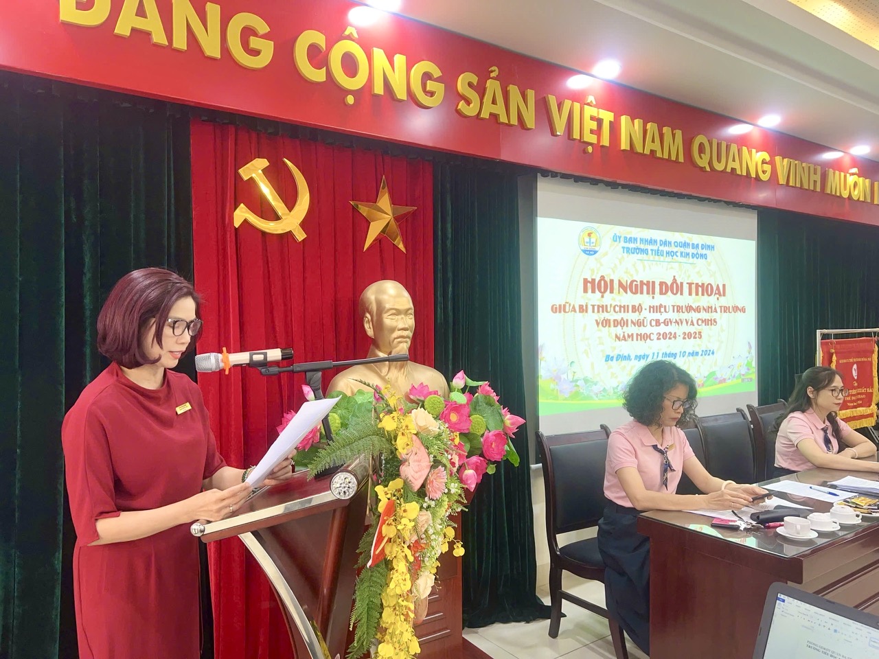 A person standing at a podium with a microphone and a person in a red dress

Description automatically generated