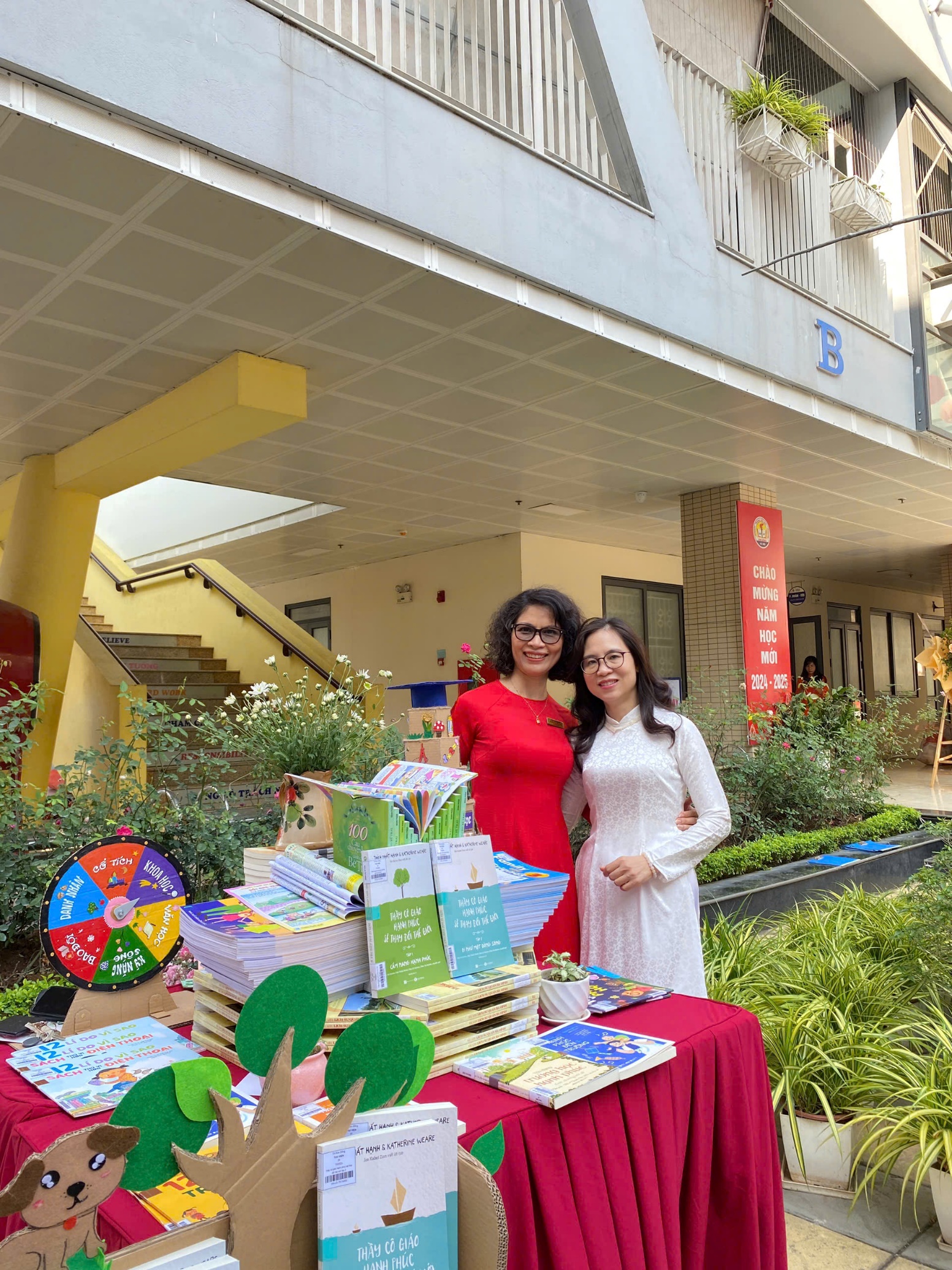 Two women standing next to a table with books and a paper treeDescription automatically generated